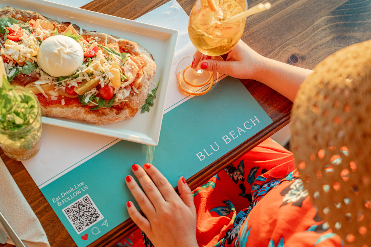 Inquadratura dall'alto di una pizza con mozzarella di bufala, con una mano femminile che ne afferra una fetta, catturata al tramonto. Fotografia realizzata da un fotografo specializzato in hotel a Pula, evidenziando l'atmosfera e la qualità culinaria del momento.