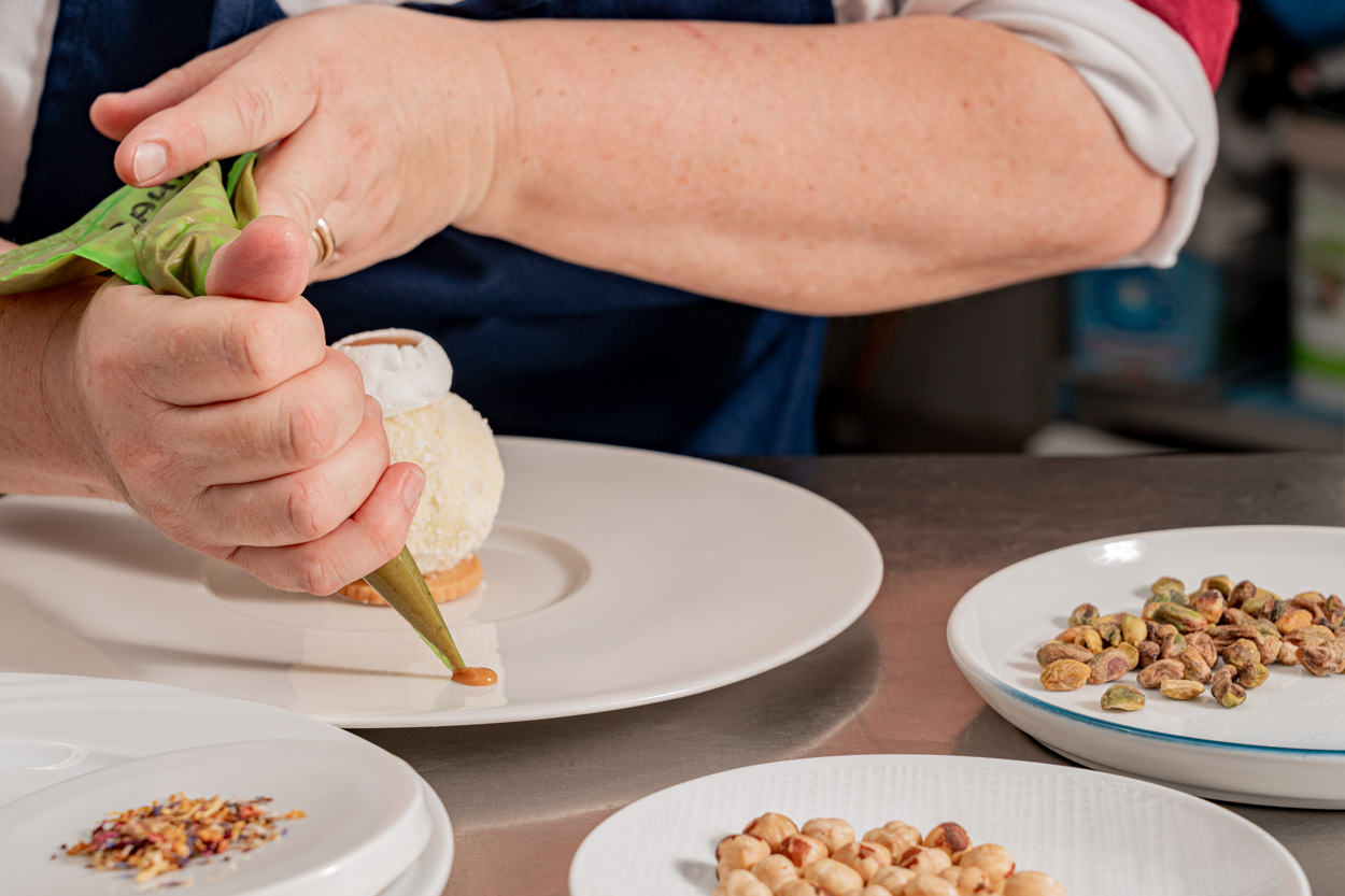 L'immagine ritrae uno chef mentre decora un dessert con una sac à poche, circondato da ingredienti di qualità come nocciole e pistacchi. Questo servizio fotografico evidenzia l'eccellenza gastronomica e l'attenzione ai dettagli dell'hotel a Porto Cervo. Ideale per promuovere l'hotel come destinazione di lusso, questa foto attrarrà clienti che cercano esperienze culinarie raffinate, migliorando l'immagine del brand e supportando le strategie di marketing.