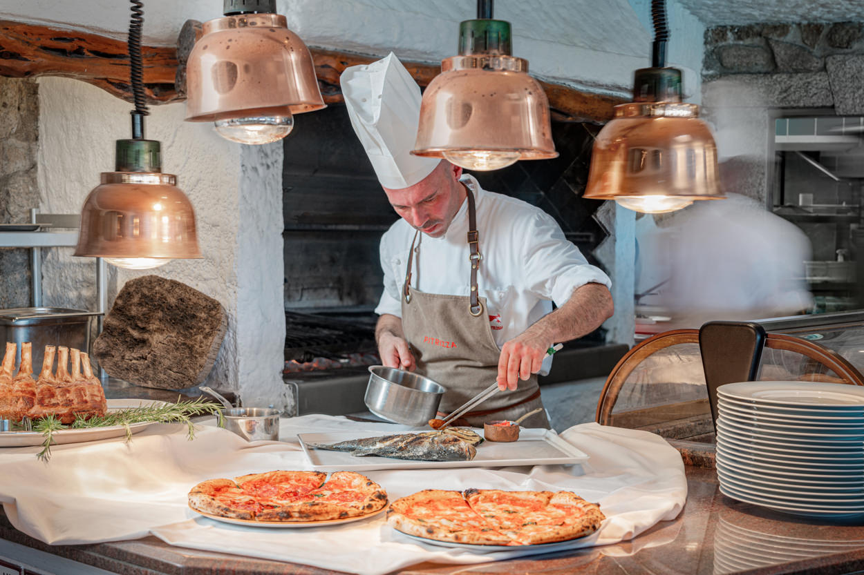 Nell'immagine del servizio fotografico per un hotel in Sardegna, uno chef è raffigurato mentre prepara un raffinato piatto di pesce in una cucina a vista, sotto la luce calda di lampade in rame. In primo piano, sono presenti anche delle pizze, rappresentando la varietà e l'eccellenza dell'offerta culinaria dell'hotel. Questa foto è ideale per promuovere l'autenticità e la qualità della cucina locale, attrarre clienti amanti della gastronomia e migliorare l'immagine del brand dell'hotel come destinazione di lusso e tradizione culinaria.