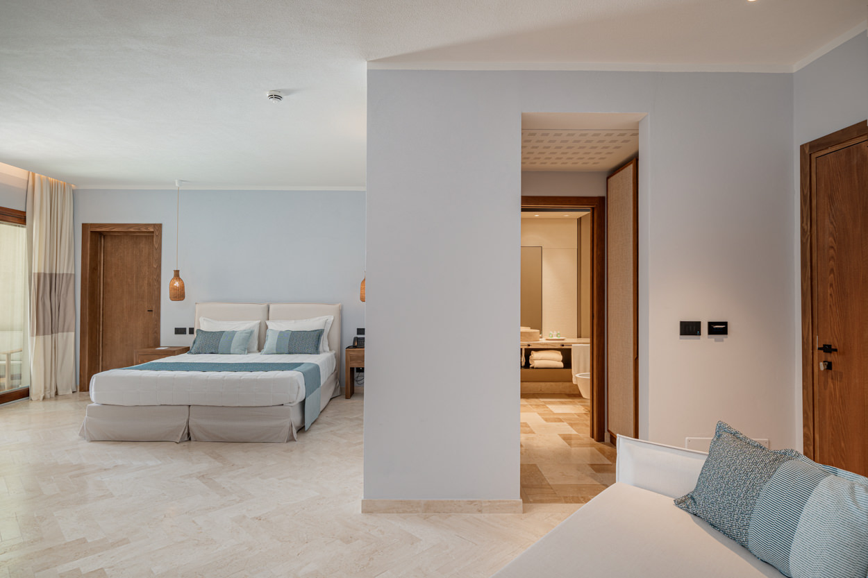 L'immagine mostra una camera da letto elegante e spaziosa di un hotel in Sardegna, con toni neutri e dettagli raffinati. L'ambiente è arredato con un letto matrimoniale, soffici cuscini decorativi, illuminazione pendente moderna e un accesso diretto al bagno, visibile sullo sfondo. Questa foto mette in risalto il design contemporaneo e il comfort della stanza, ideale per promuovere l'hotel come una destinazione di lusso e relax. Perfetta per attirare clienti che cercano un'esperienza di soggiorno raffinata, migliorando l'attrattività del brand e supportando strategie di marketing per il settore dell'ospitalità di alta gamma.